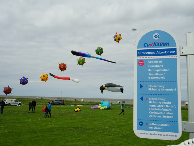 Das Bild zeigt die Drachenflugtage im Jahr 2022 in Altenbruch. Drachen in verschiedenen Farben und Formen sind am Himmel zu sehen. Links sieht man ein Schild vom Nordseeheilbad Cuxhaven, das als Wegweiser dient.