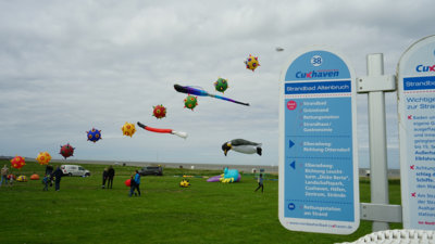 Das Bild zeigt die Drachenflugtage im Jahr 2022 in Altenbruch. Drachen in verschiedenen Farben und Formen sind am Himmel zu sehen. Links sieht man ein Schild vom Nordseeheilbad Cuxhaven, das als Wegweiser dient.