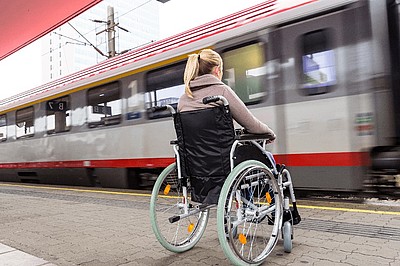 Frau im Rollstuhl am Bahnhof mit vorbeifahrendem Zug