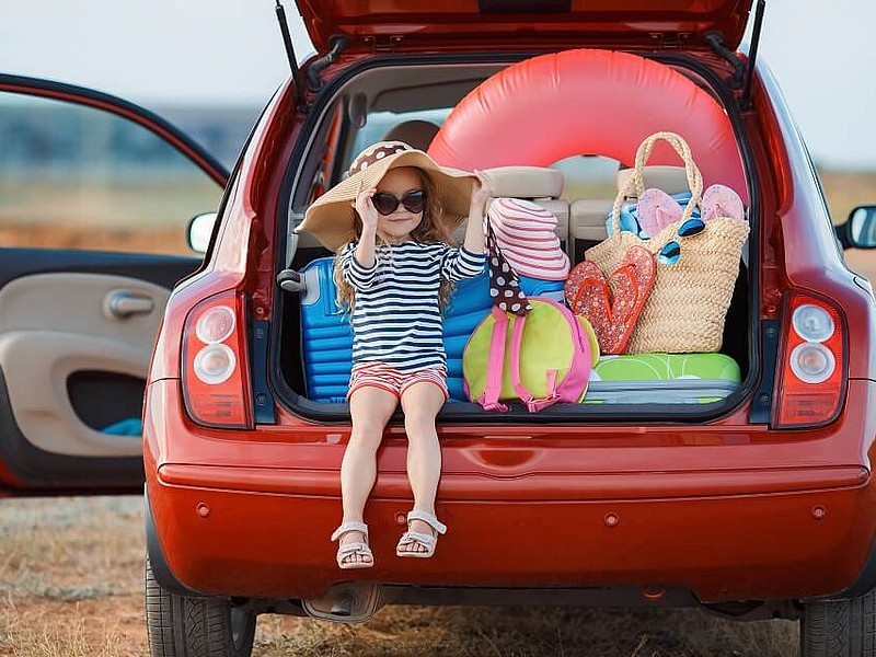 Rotes Auto mit offenem Kofferraum, in dem ein Kind mit unterschiedlichen Koffern sitzt