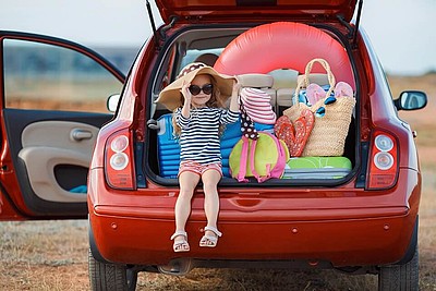Rotes Auto mit offenem Kofferraum, in dem ein Kind mit unterschiedlichen Koffern sitzt